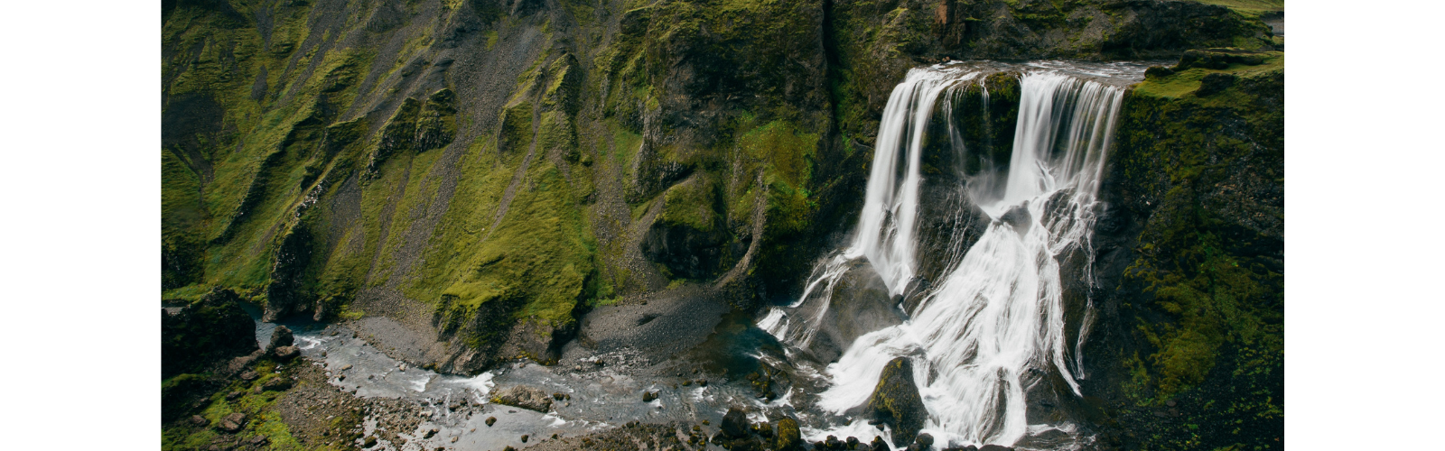 A tall waterfall crashes over large rocks below, flowing into a river. The waterfall is surrounded by lush, green cliffs. 